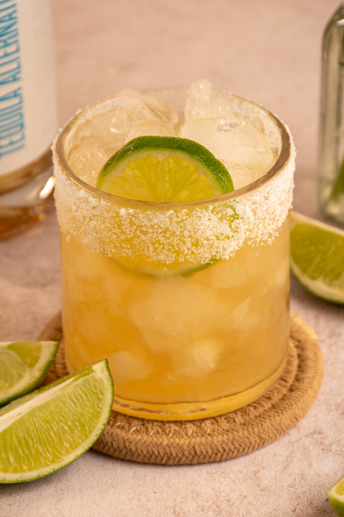 A close up of a glass with a salted rim filled with ice and ritual zero proof margarita with a lime slice.