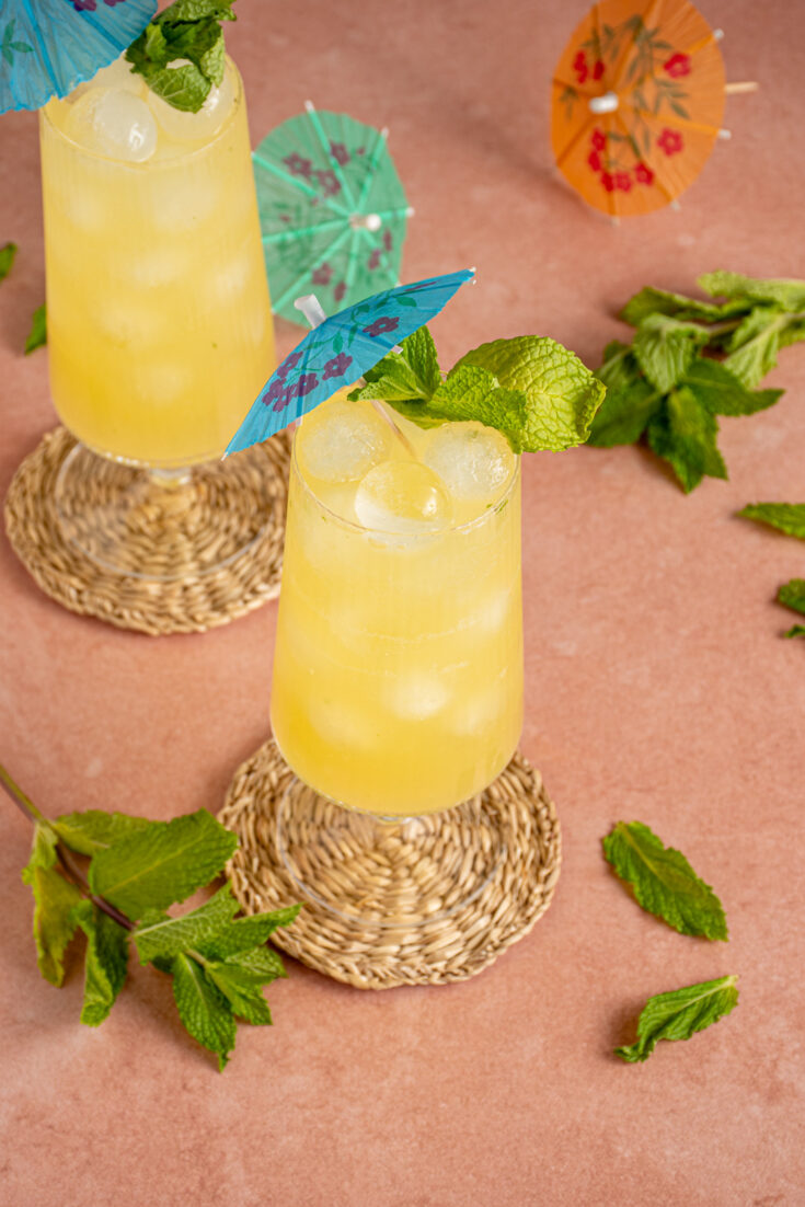A glass with yellow colored mint pineapple ginger mocktail garnished with a fresh sprig of mint and a blue drink umbrella.