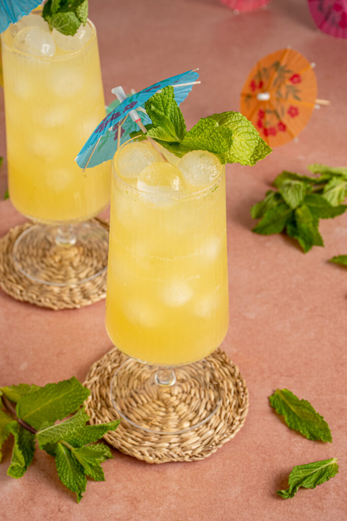 A glass with yellow colored mint pineapple ginger mocktail garnished with a fresh sprig of mint and a blue drink umbrella.