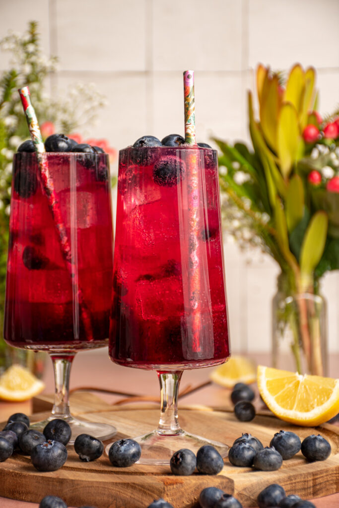 A pair of glasses with brightly colored blueberry ginger beer mocktail garnished with fresh blueberries.