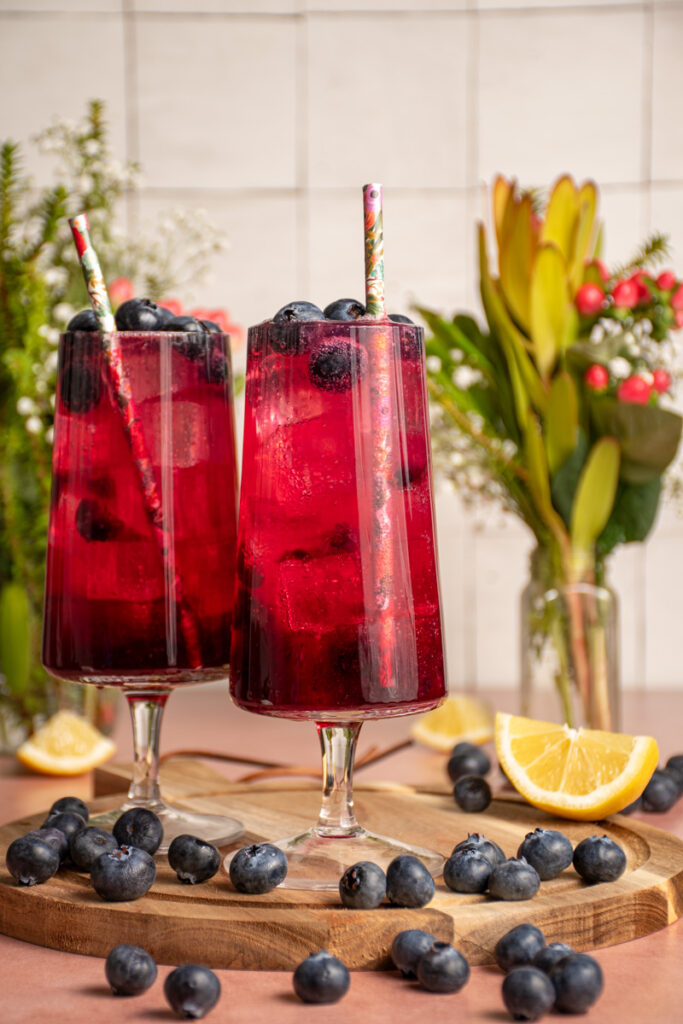 A pair of glasses with brightly colored blueberry ginger beer mocktail garnished with fresh blueberries.