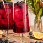 A pair of glasses with brightly colored blueberry ginger beer mocktail garnished with fresh blueberries.