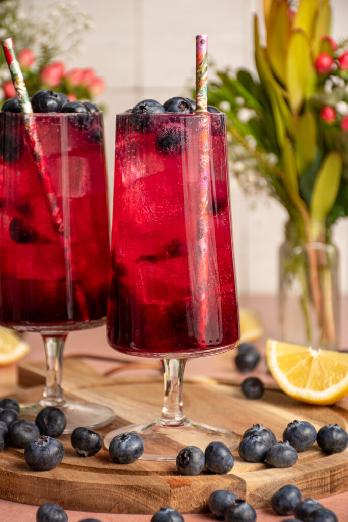 A pair of glasses with brightly colored blueberry ginger beer mocktail garnished with fresh blueberries.