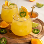A glass with yellow colored Jalapeño Orange Margarita garnished with orange slice, lime slice, and jalapeno slice.