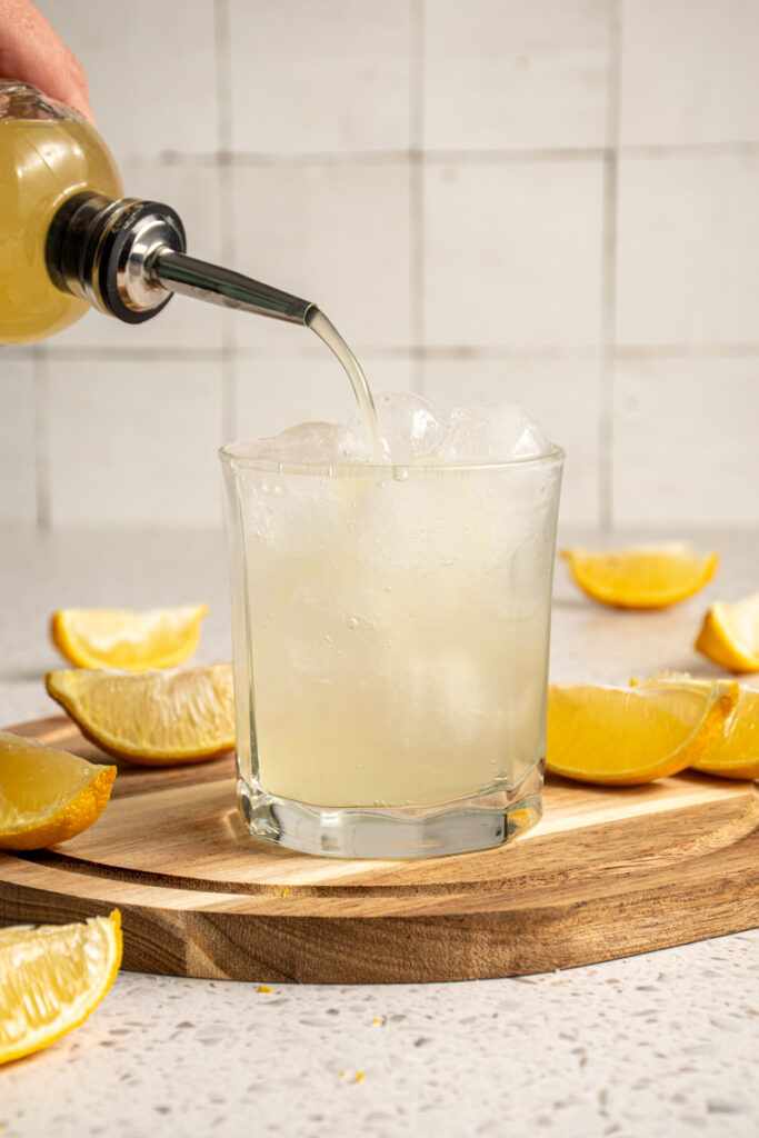 Pouring lemon ginger shrub into a glass of sparkling water.