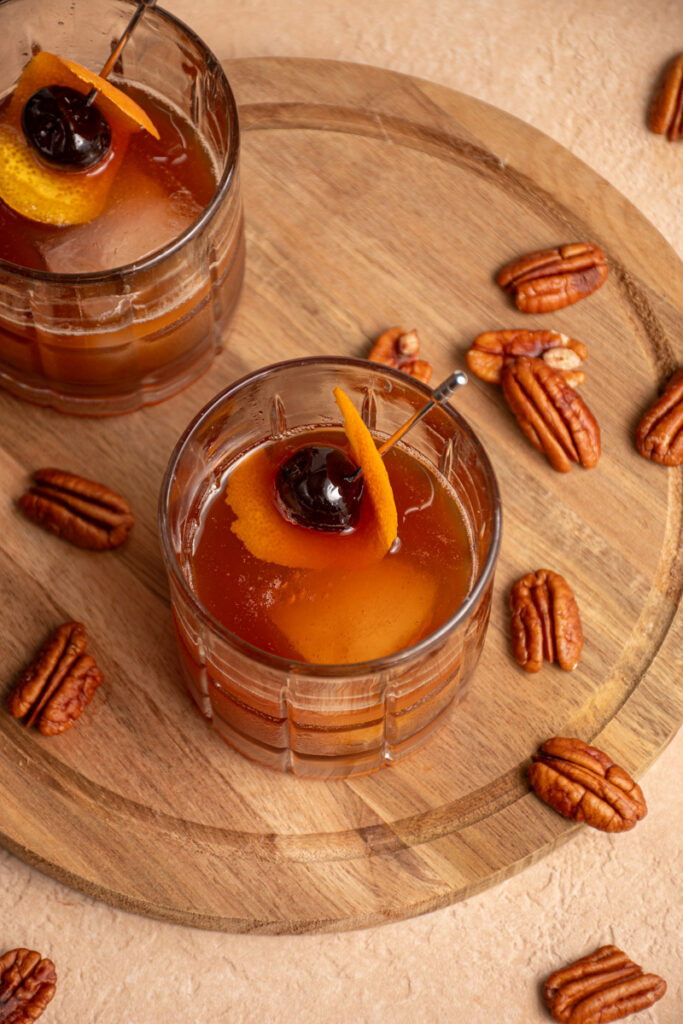 A glass filled with ice and a pecan old fashioned garnished with a cherry and orange peel.
