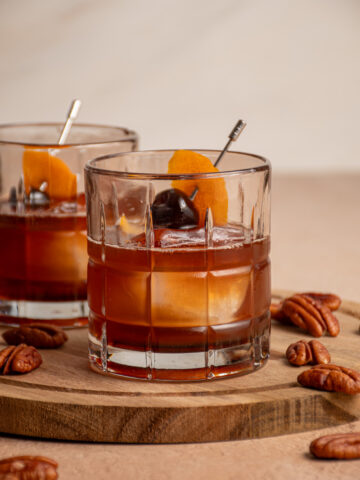 A glass filled with ice and a pecan old fashioned garnished with a cherry and orange peel.